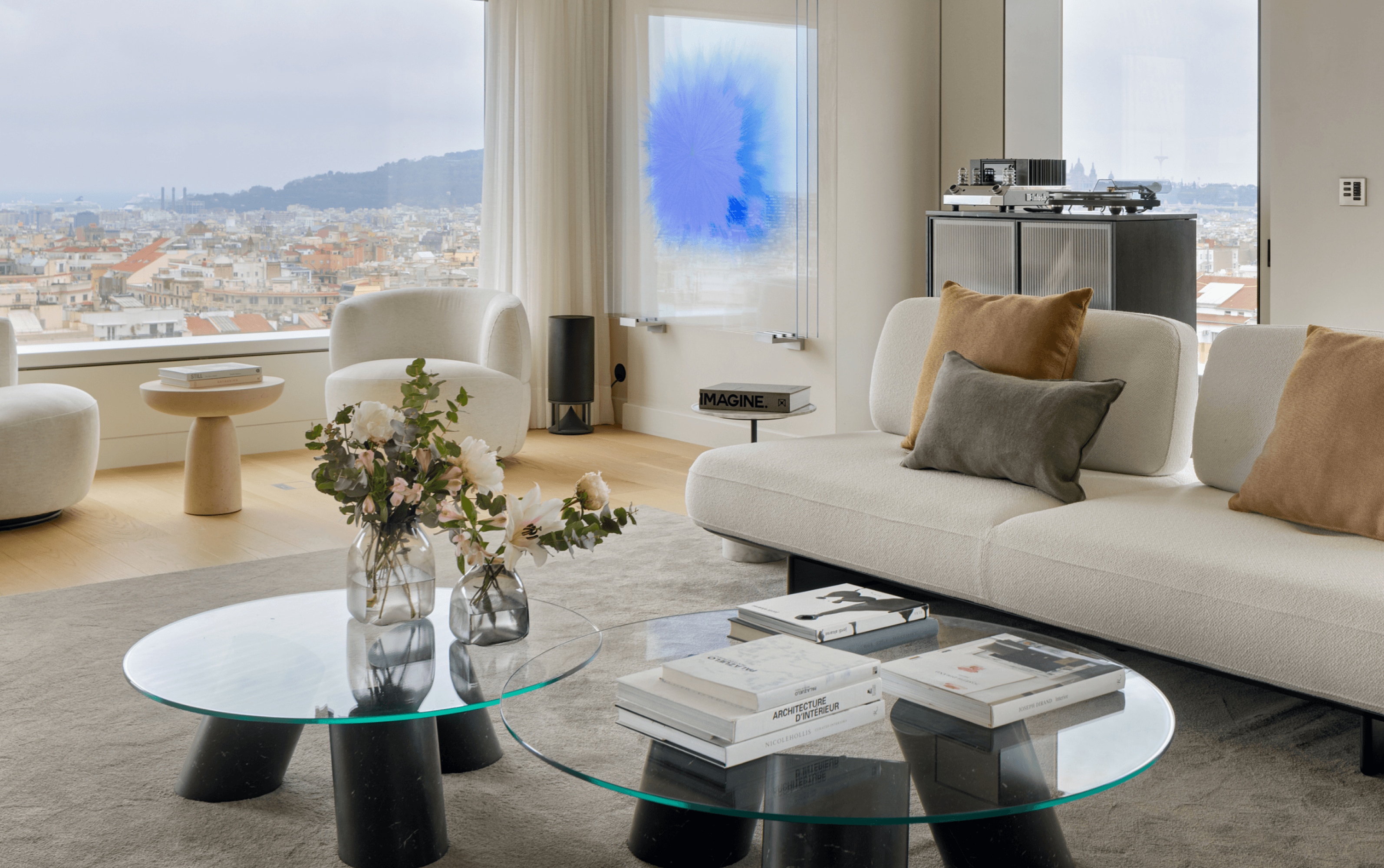 Bright living room with panoramic city views, white sofas, round glass tables, and contemporary artwork in soft blue tones.