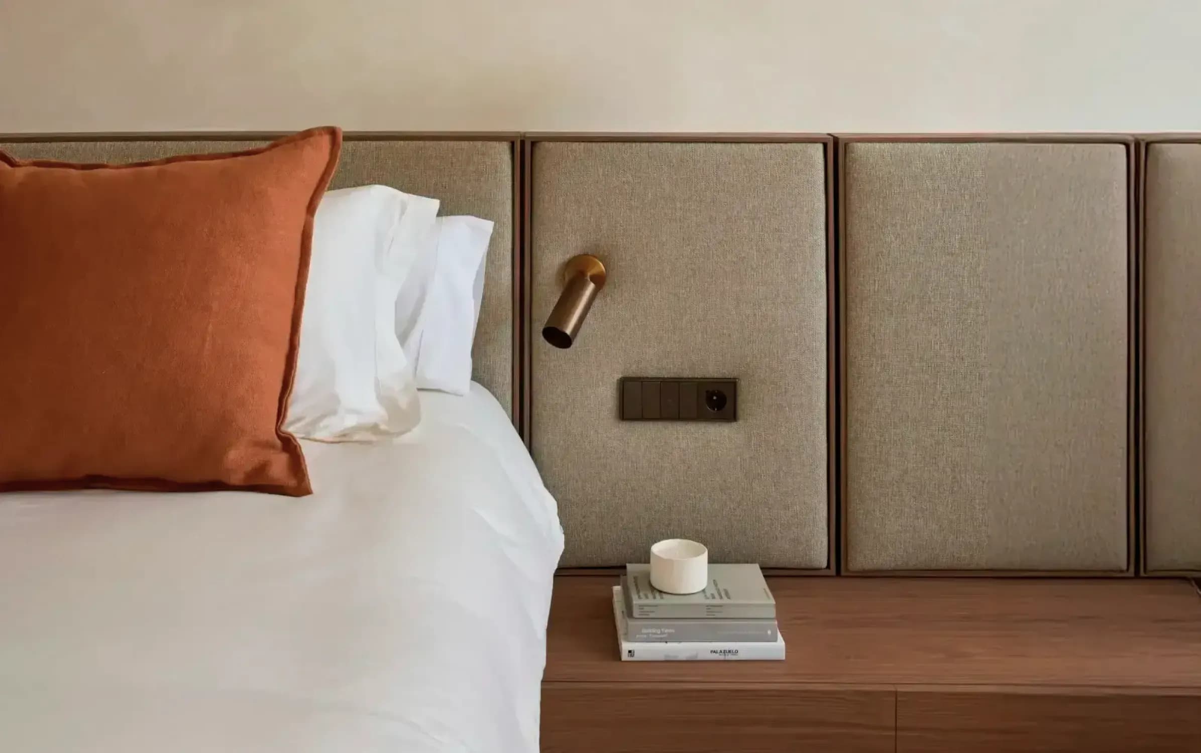 Minimalist bedroom with large orange cushion, padded headboard, brass wall light, and books on a wooden nightstand.